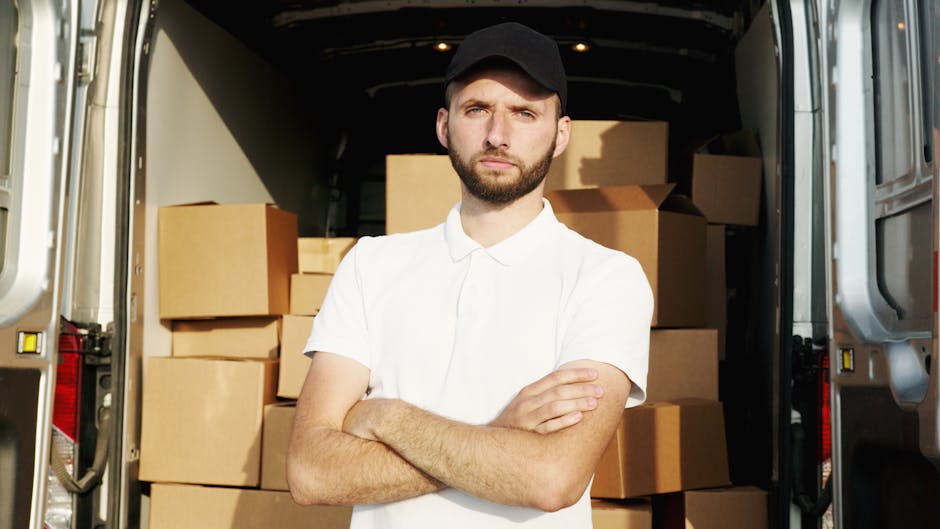 A man arranging cardboard boxes outside a white moving van parked on a residential street with houses and greenery in the background, preparing for a home relocation. The man is holding a box, and sev